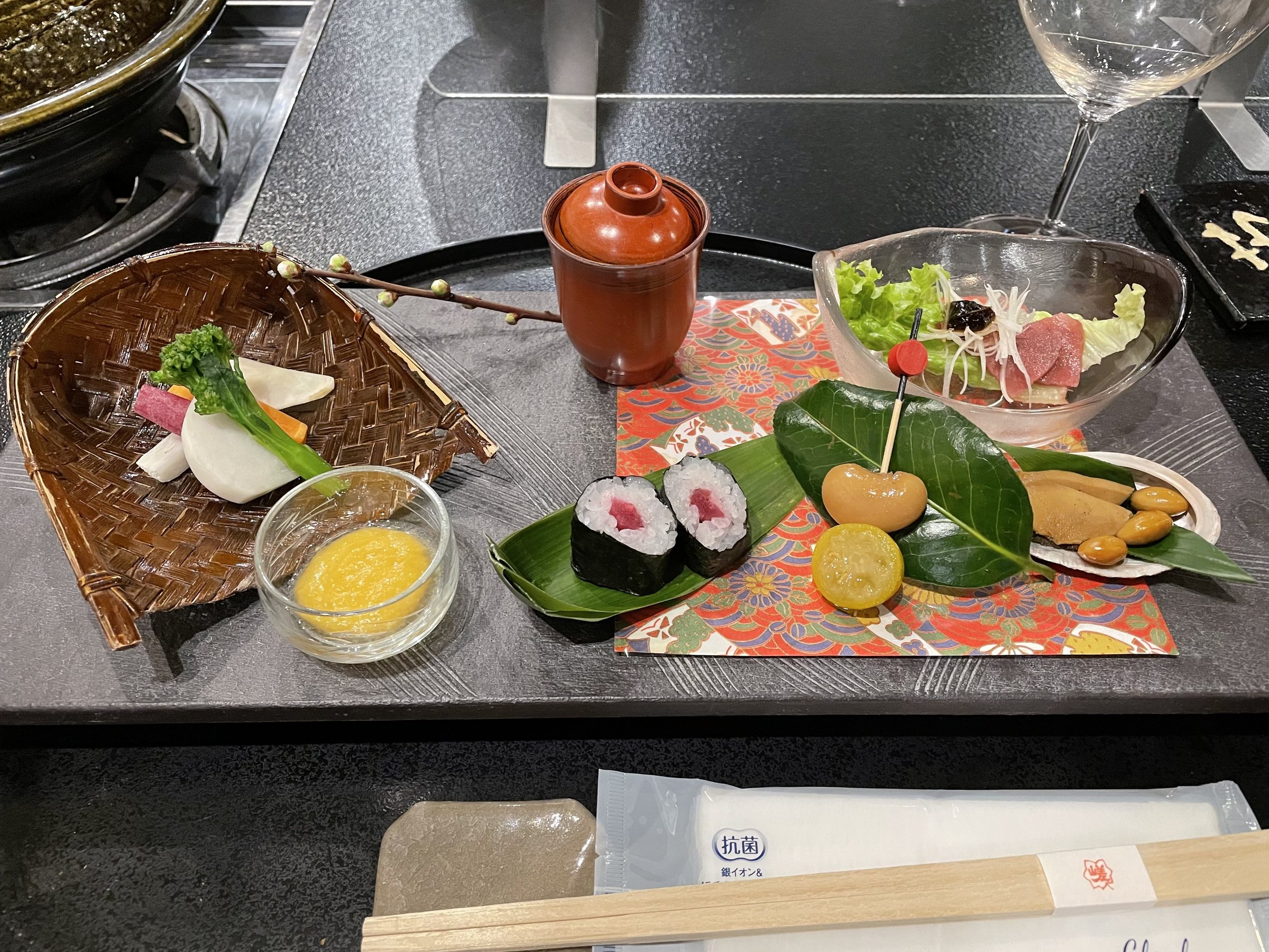The dinner table set with the appetiser plate: vegetables in a woven basket, rolled sushi and small skewered bites, with a wine glass waiting.