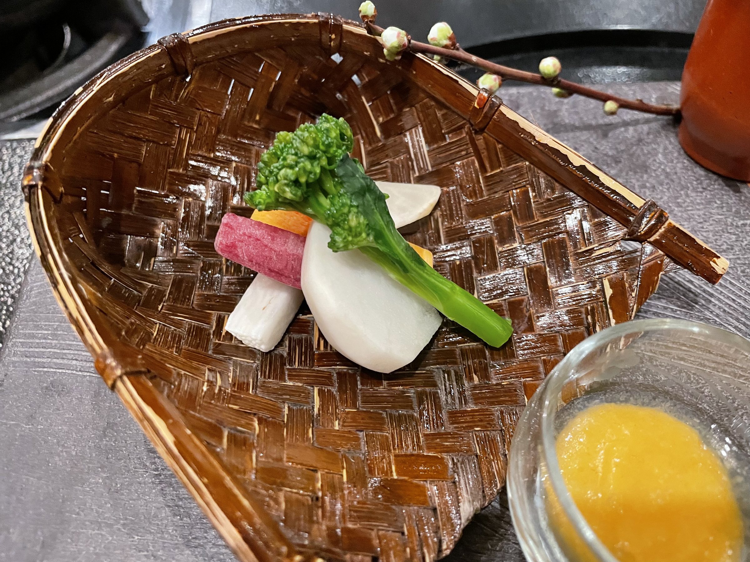 Local vegetables, including rape blossom and radish, in a woven bamboo basket beside a glass of dip.