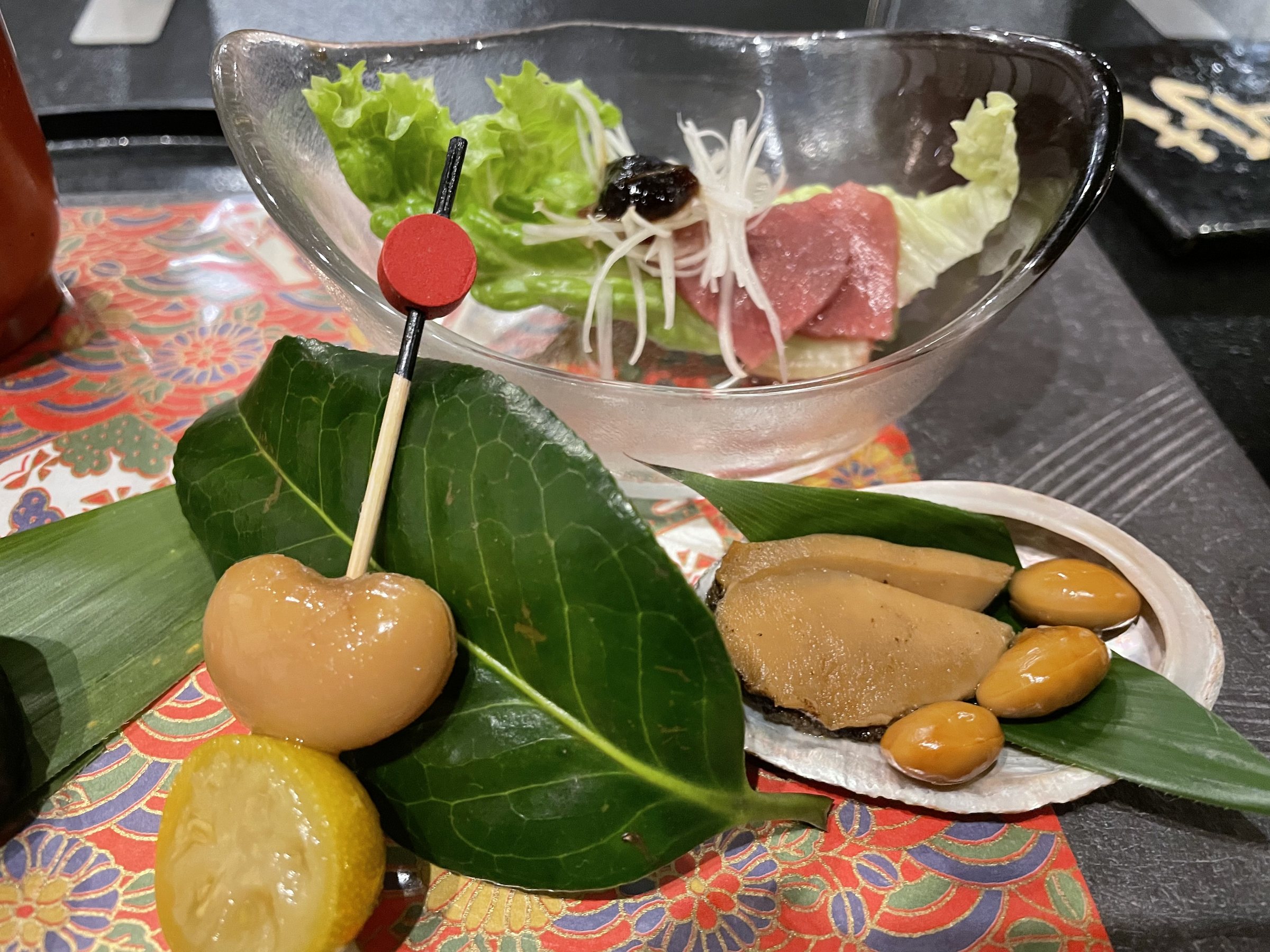 Small skewered bites and simmered morsels on leaves, with a salad in a glass bowl behind.