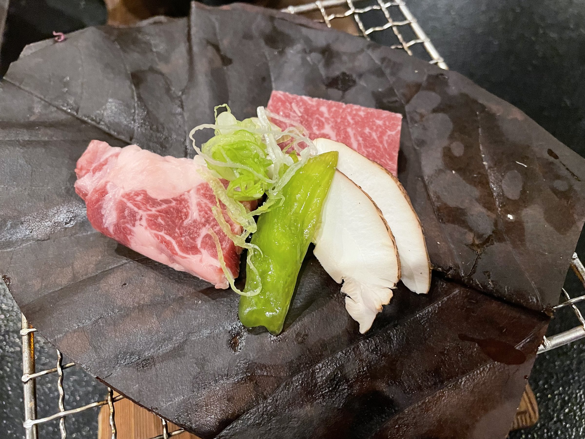 Marbled beef, a green pepper and sliced mushroom on a dried leaf over a small grill.