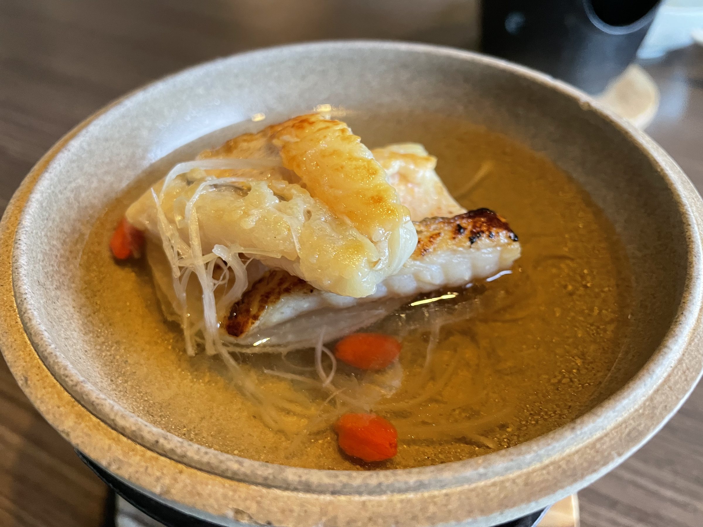 Tilefish with its scales crisped, in broth in a stoneware pot over a burner.
