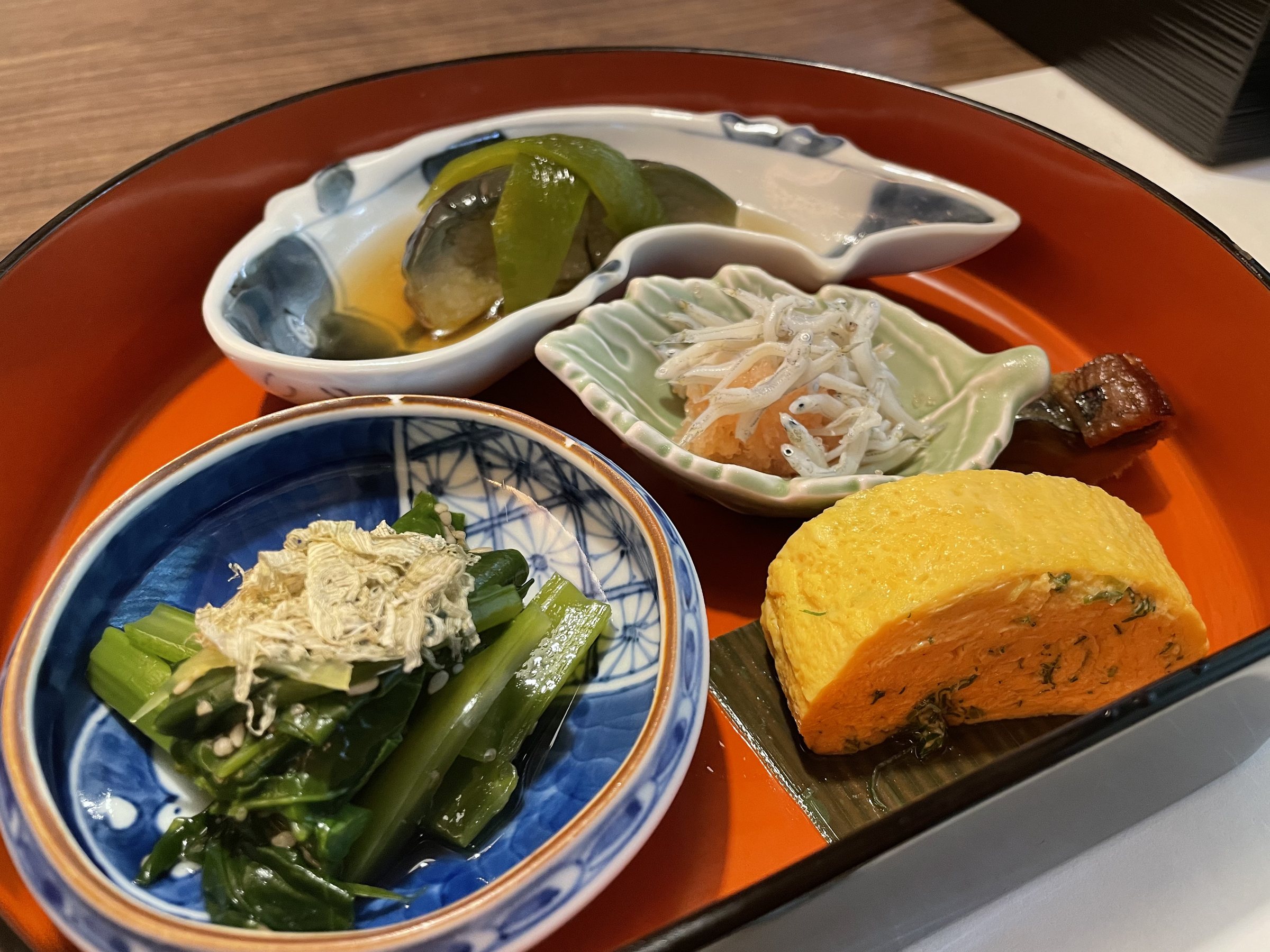 Small breakfast dishes on a red lacquered tray, including greens with whitebait and rolled omelet.