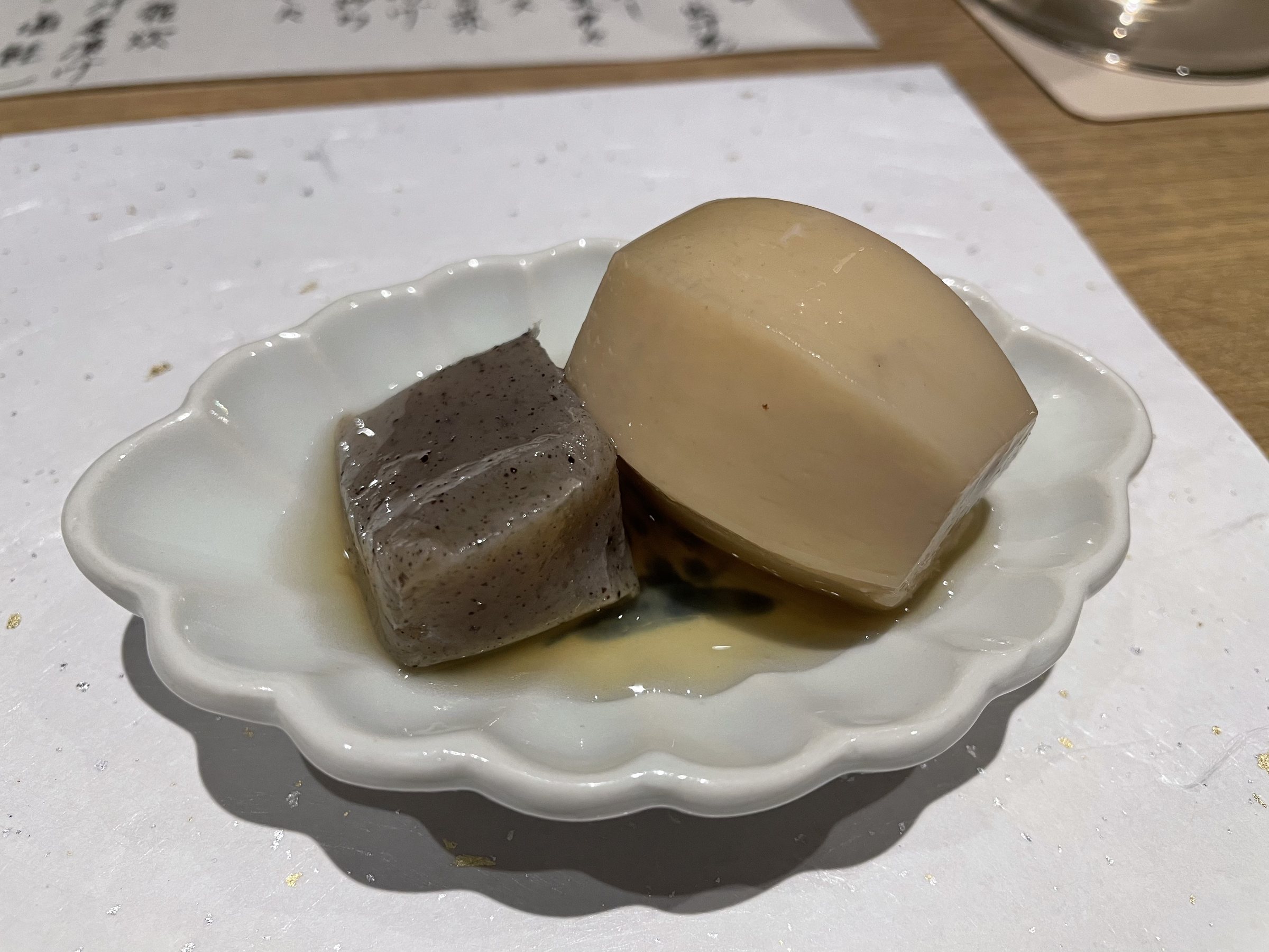 A block of konnyaku and a thick piece of simmered daikon on a scalloped plate.