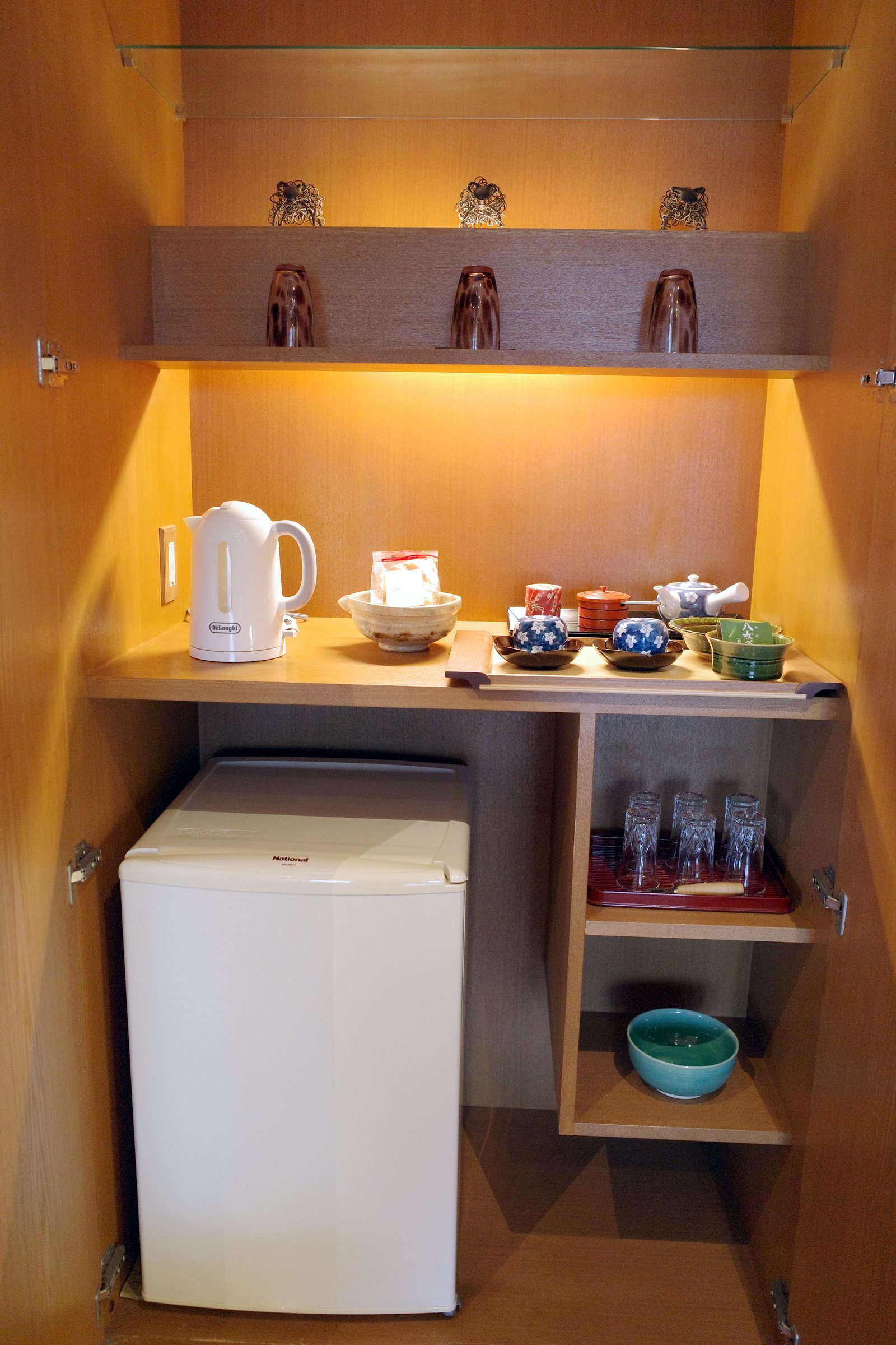 The tea station in a lit cupboard: kettle, tea set and a small fridge.