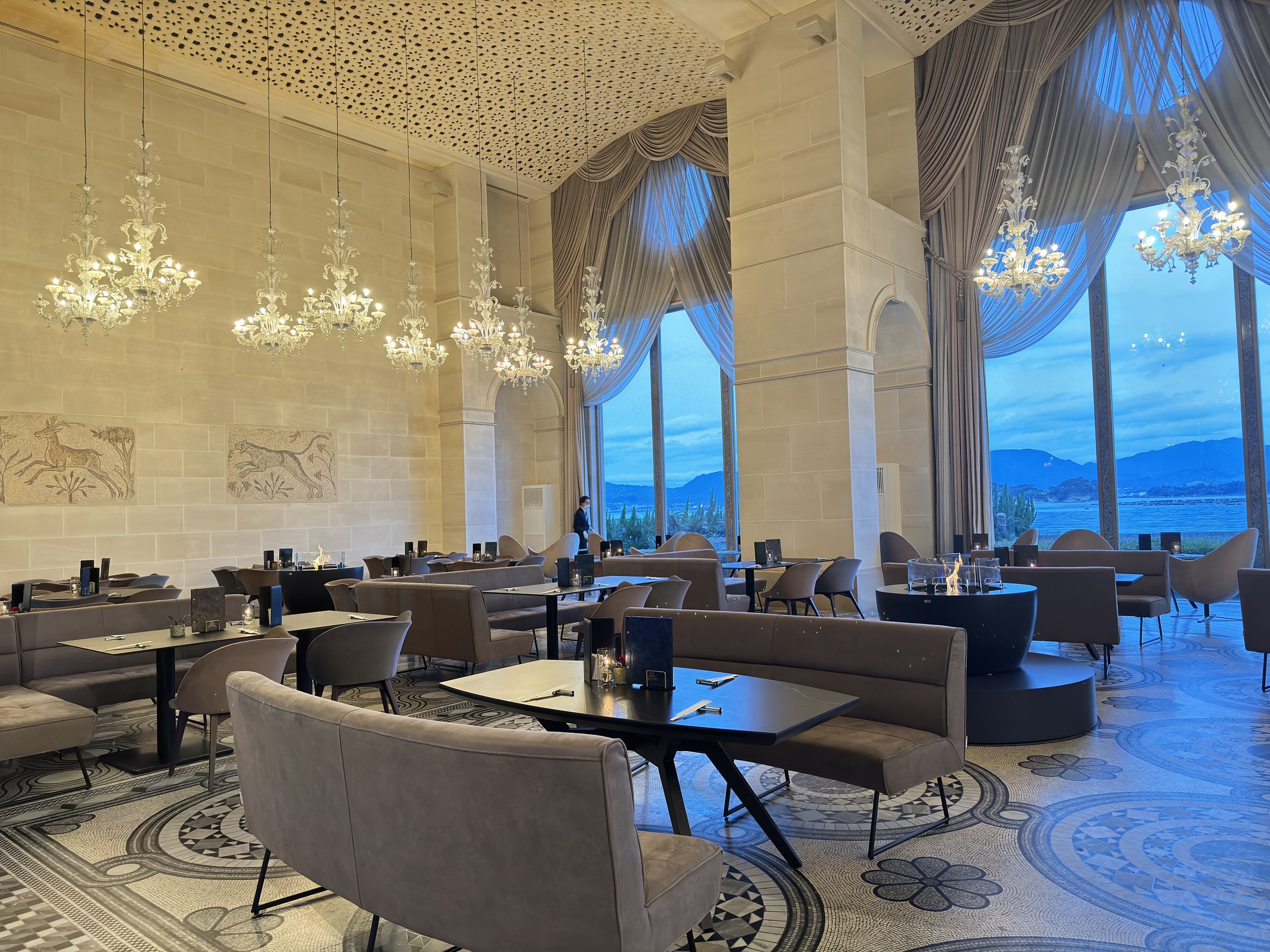 Glass chandeliers hang before the arched windows, with the sea and mountains beyond.