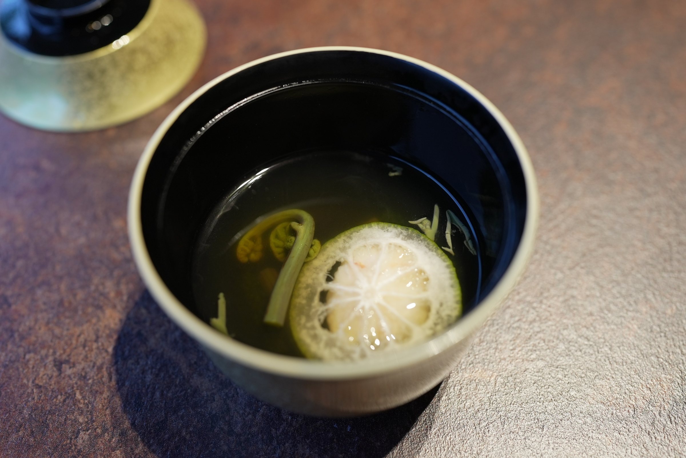 The clear soup, lid lifted: a pale dumpling and spring greens in golden dashi.