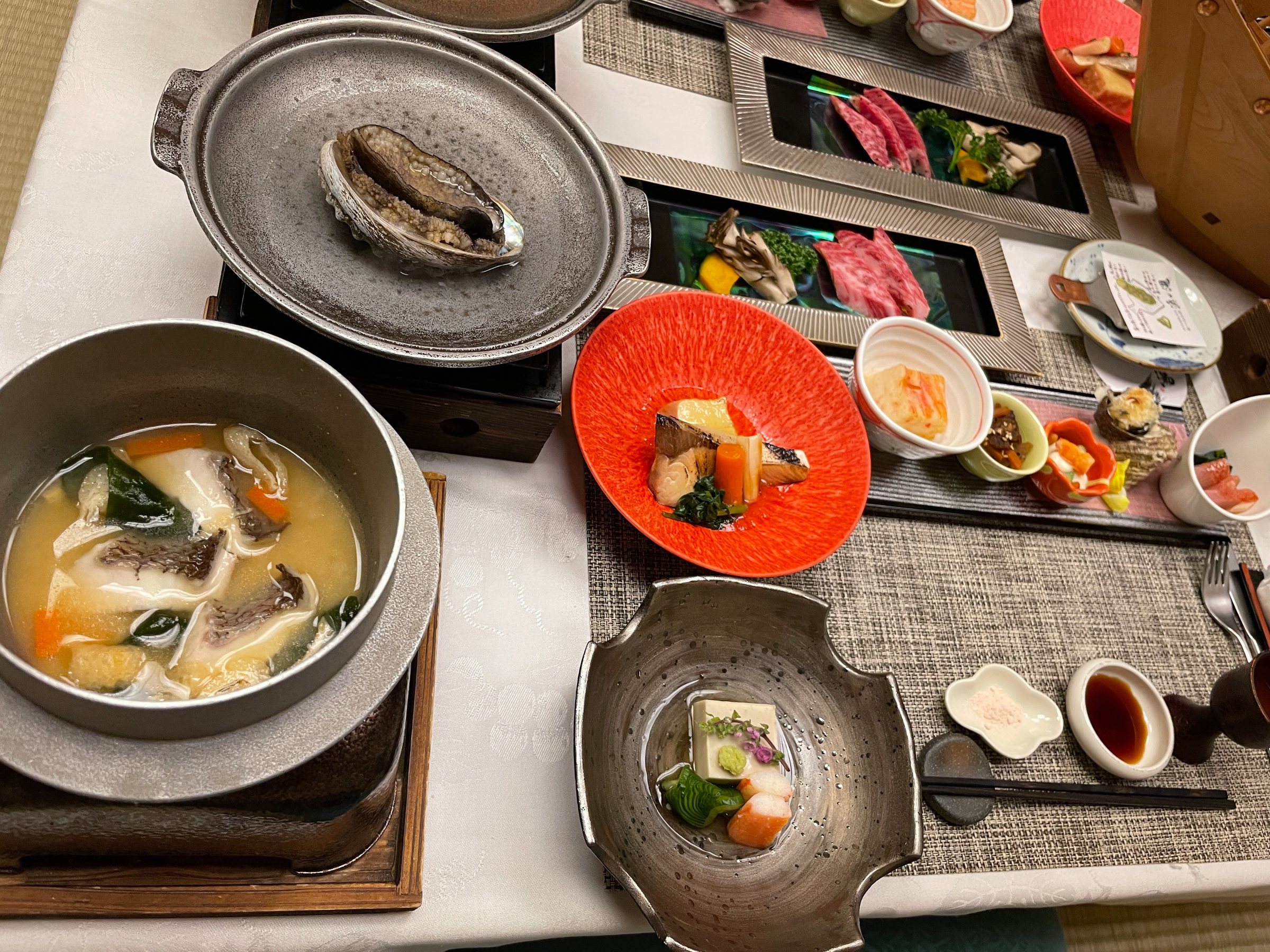 Abalone in its shell over a burner, beside a seafood soup and a simmered dish.