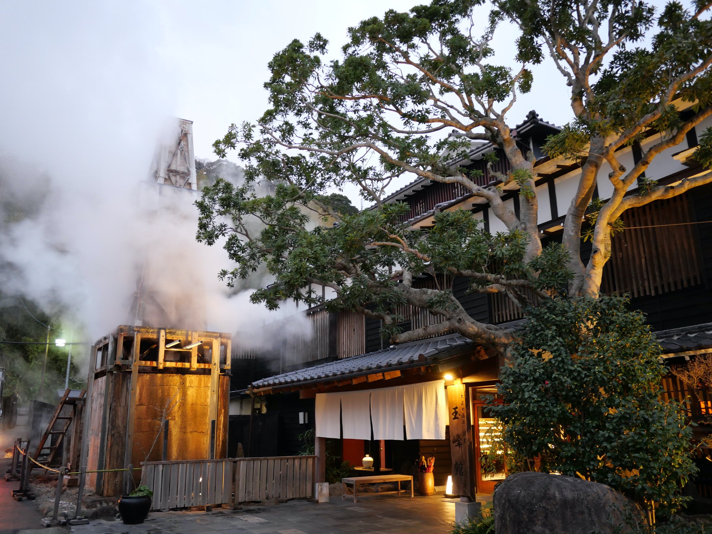 Dusk at the entrance: steam billows from the timber source tower beside the white noren.