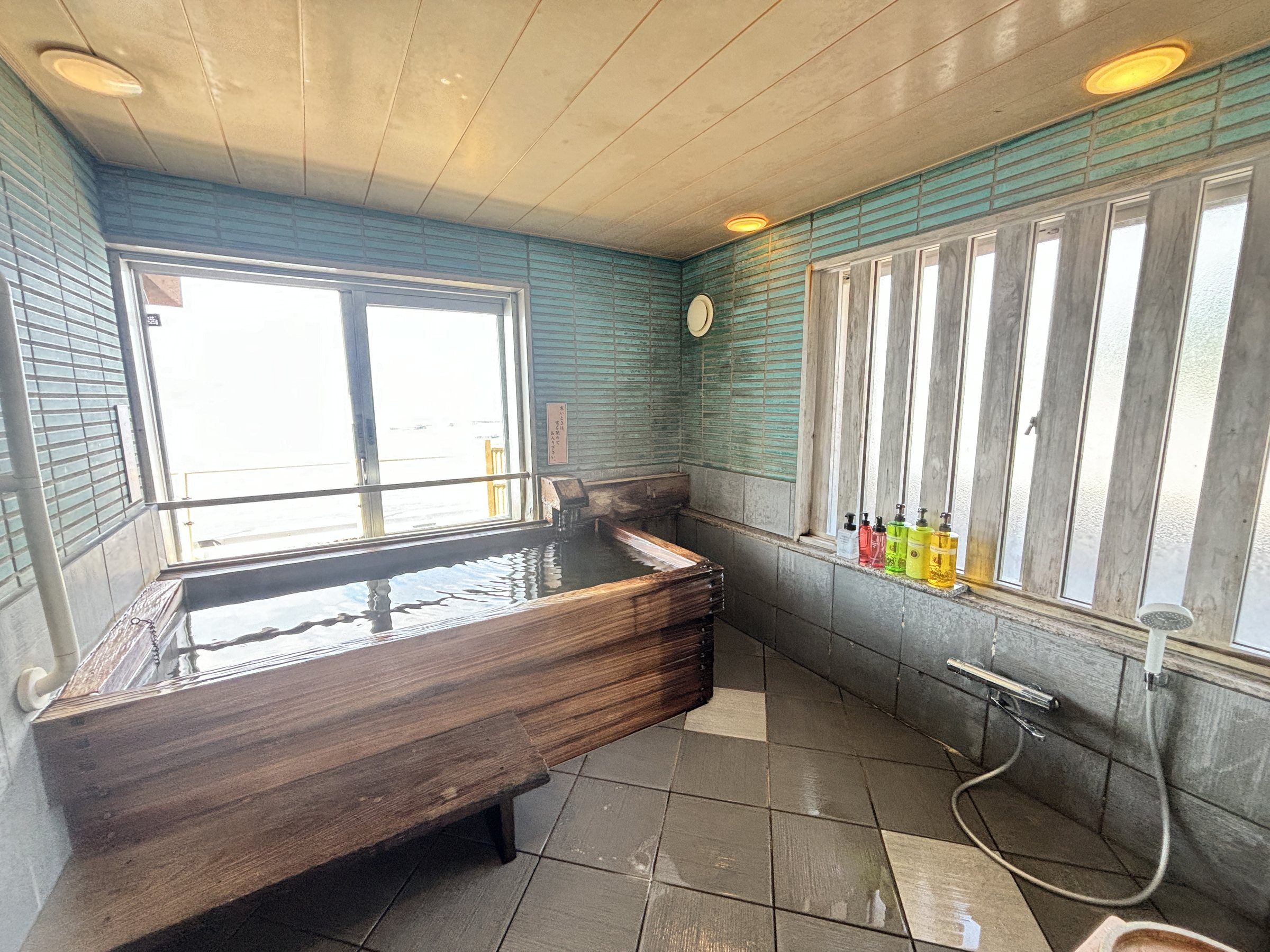 A wooden tub in a tiled bathroom, with windows onto the sea.