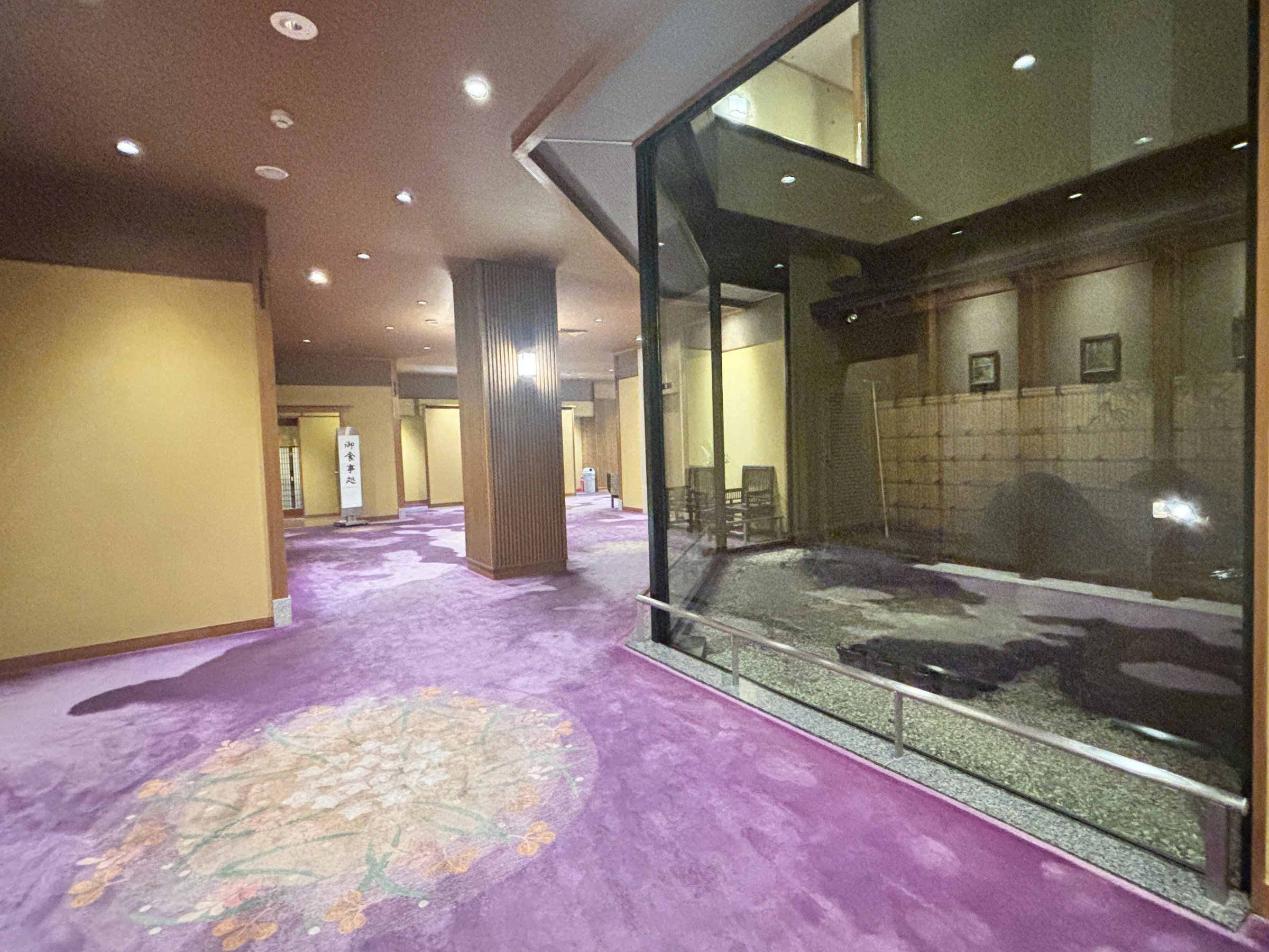 A wide, bright corridor with a patterned purple carpet and a glass-walled courtyard.