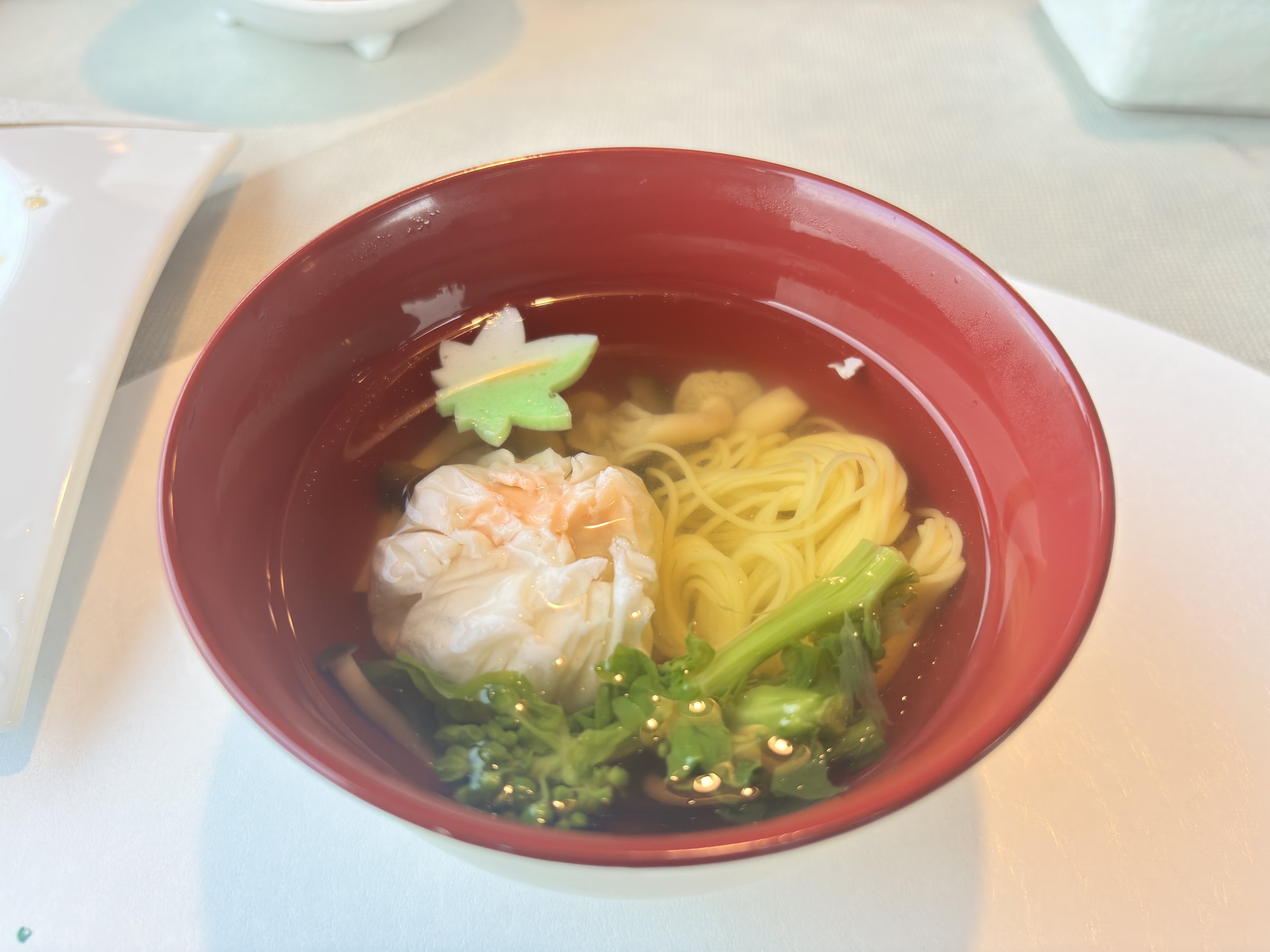 A red bowl of clear broth with pale yellow noodles, a shinjo dumpling and greens.