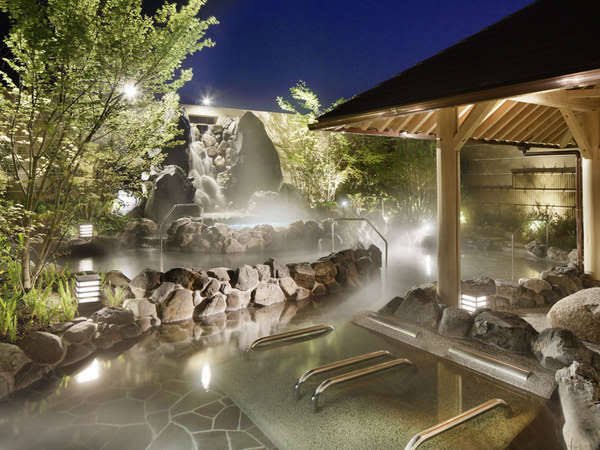 The rock-lined open-air bath at night, among trees and steam. (Photo: Kobe Minato Onsen Ren official website)