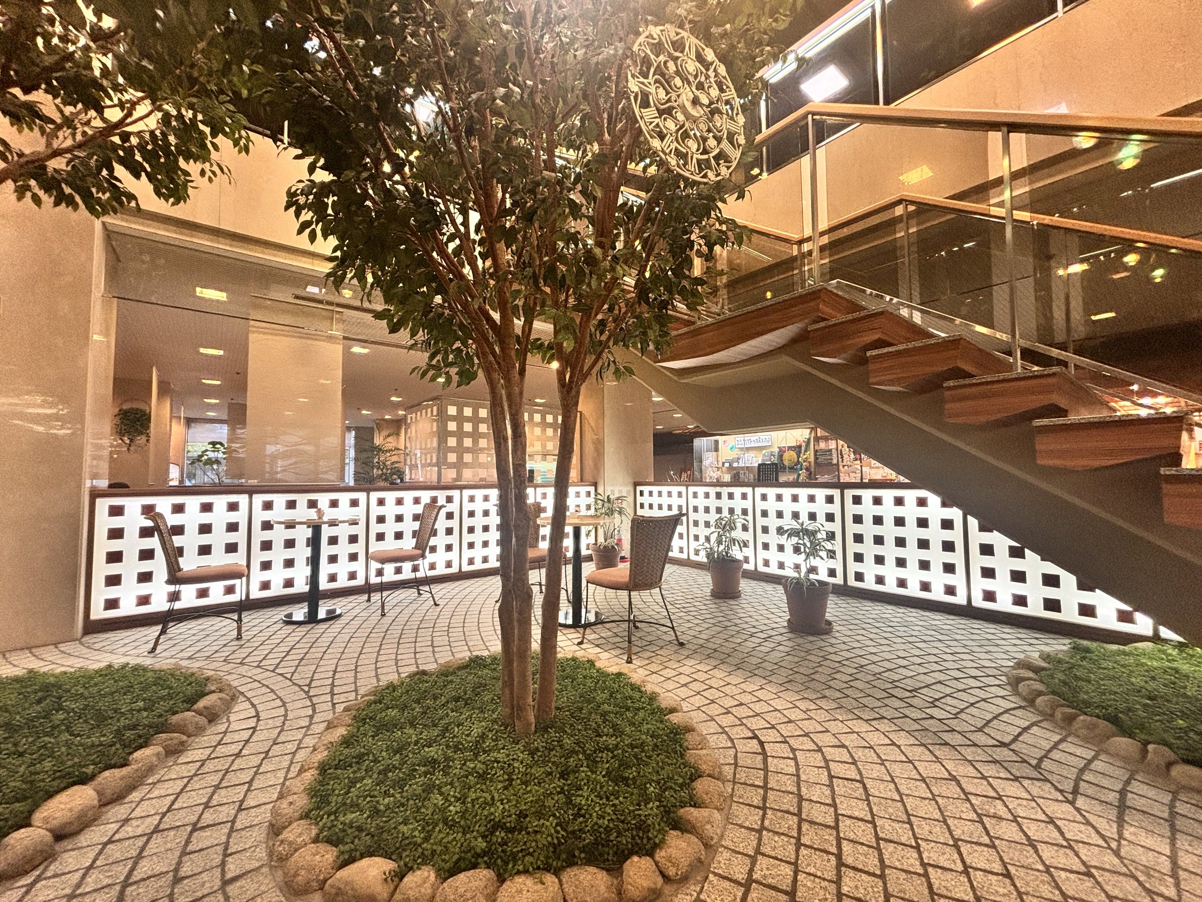 A tree in a stone-paved atrium beneath an open staircase.