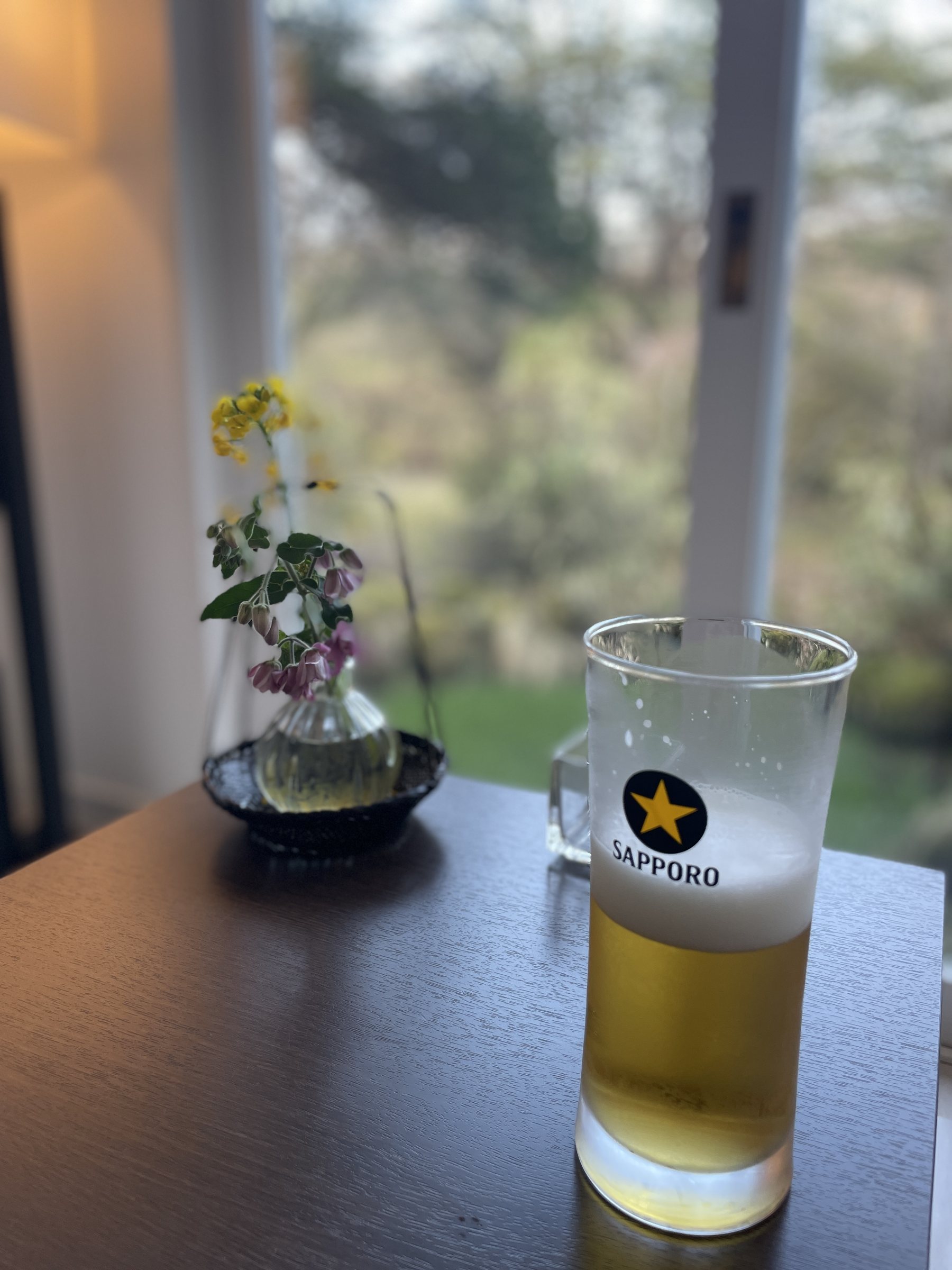 A glass of beer by the window, beside a small vase of spring flowers.