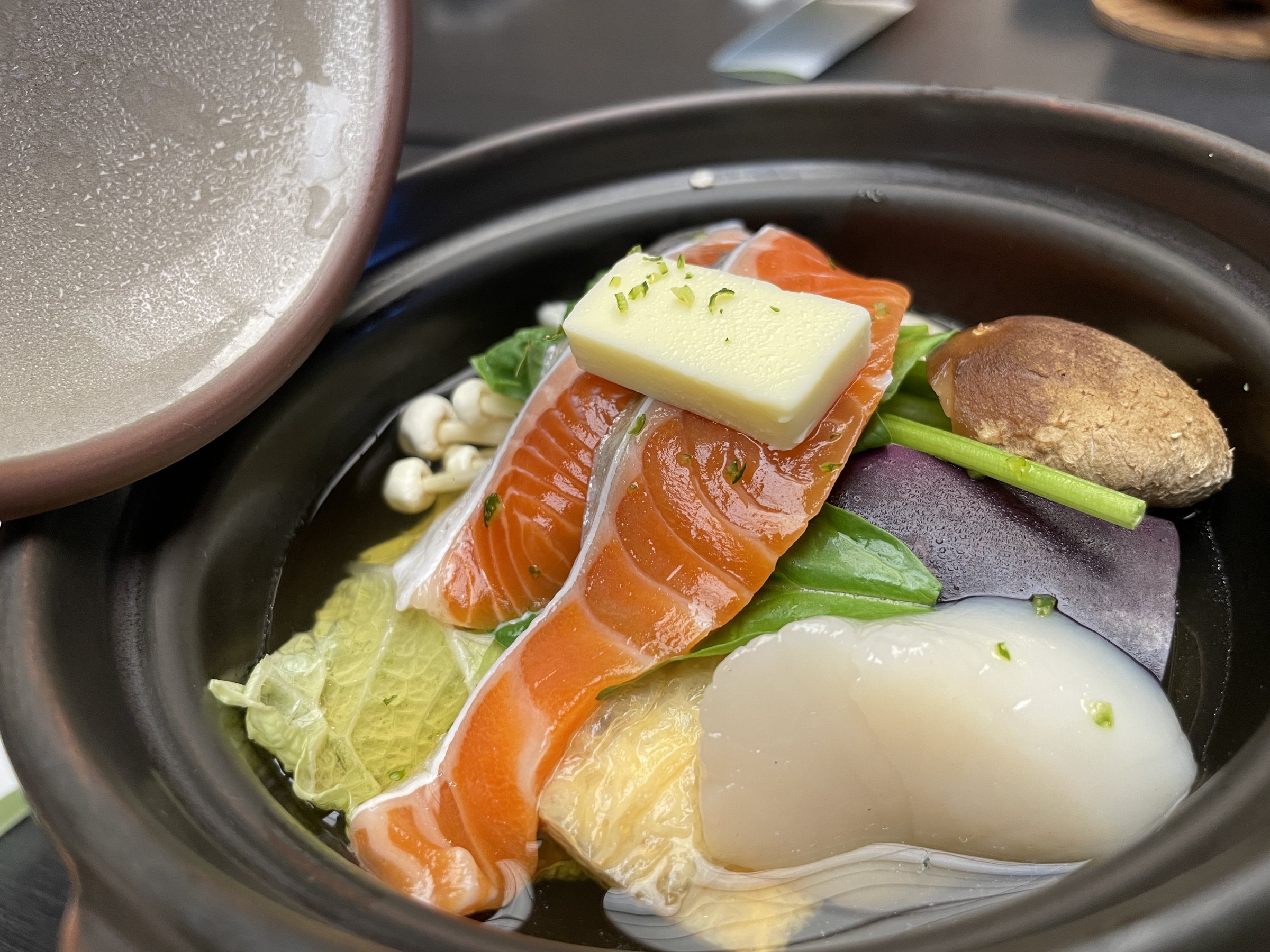 A hot pot of salmon and a large scallop, topped with a pat of butter.
