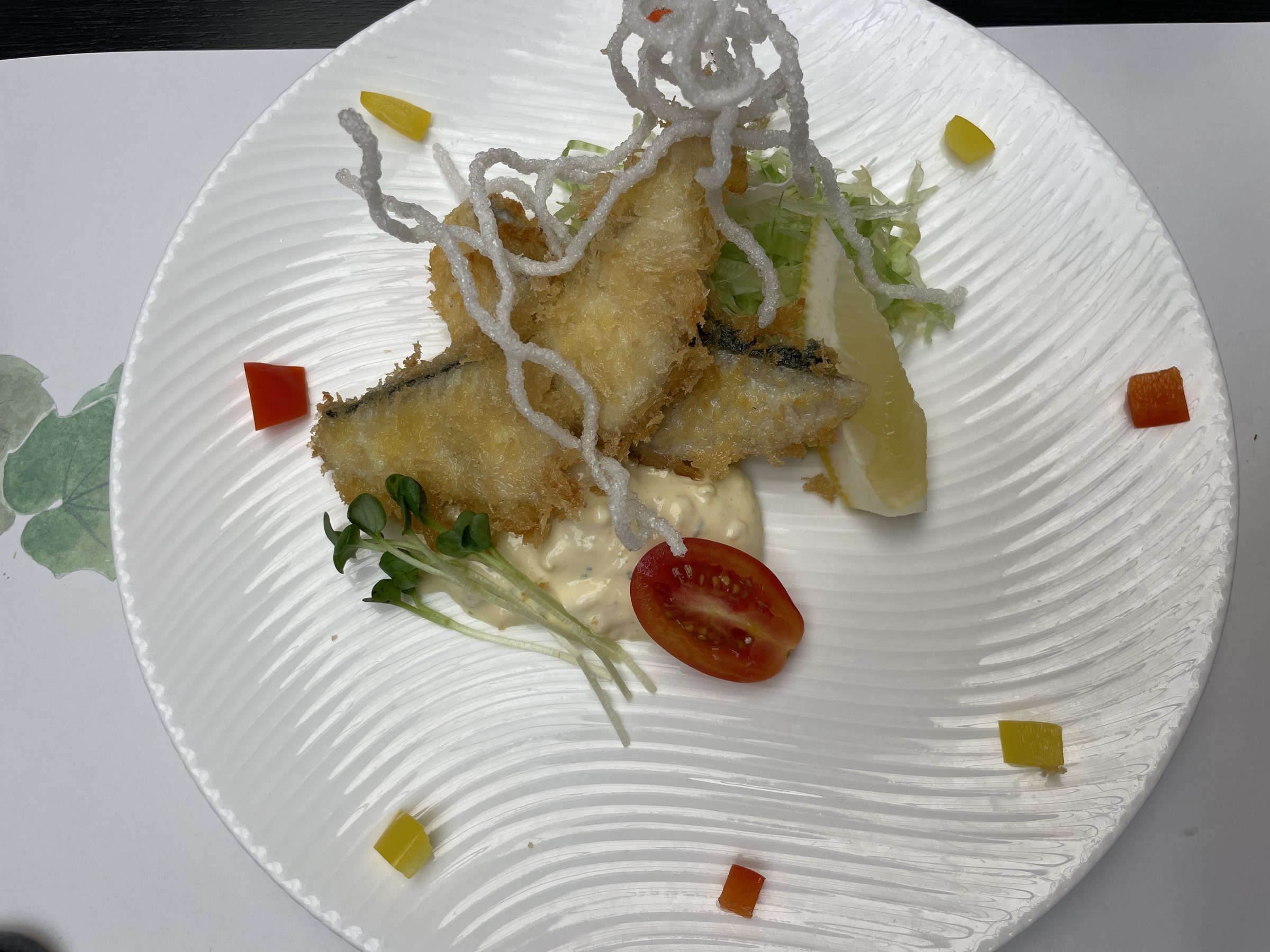 Fried fish with tartar sauce, a cherry tomato and crisp noodle strands on a white plate.