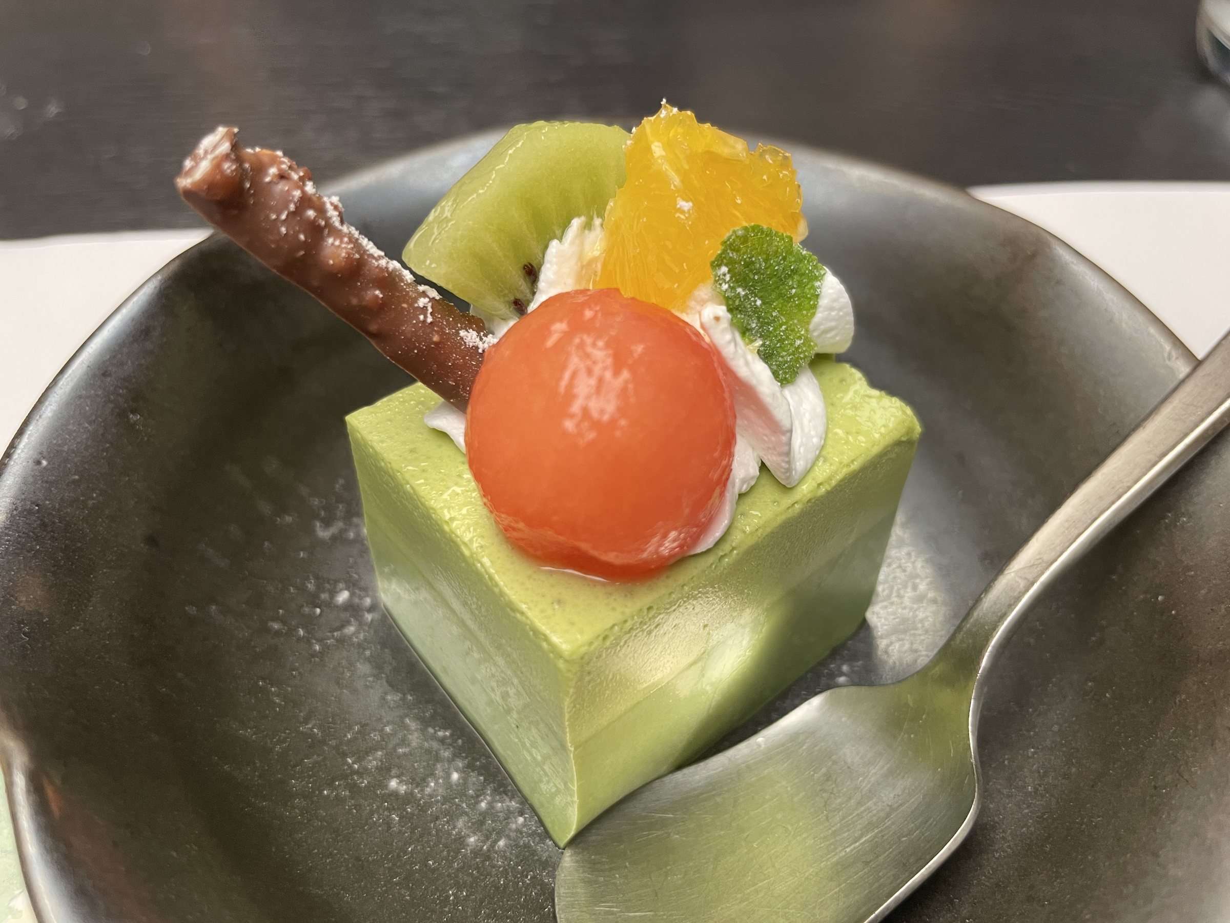 A green matcha panna cotta topped with fruit and a chocolate stick.