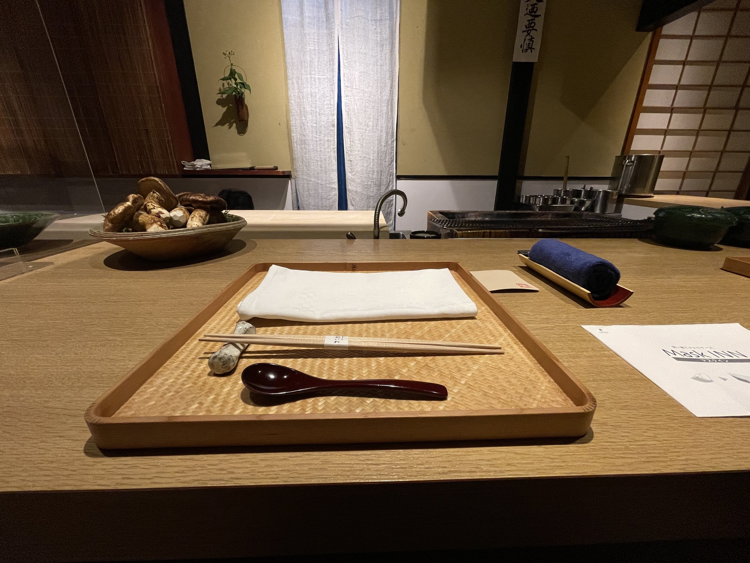 A place setting at the counter, with the kitchen and its noren curtains beyond.