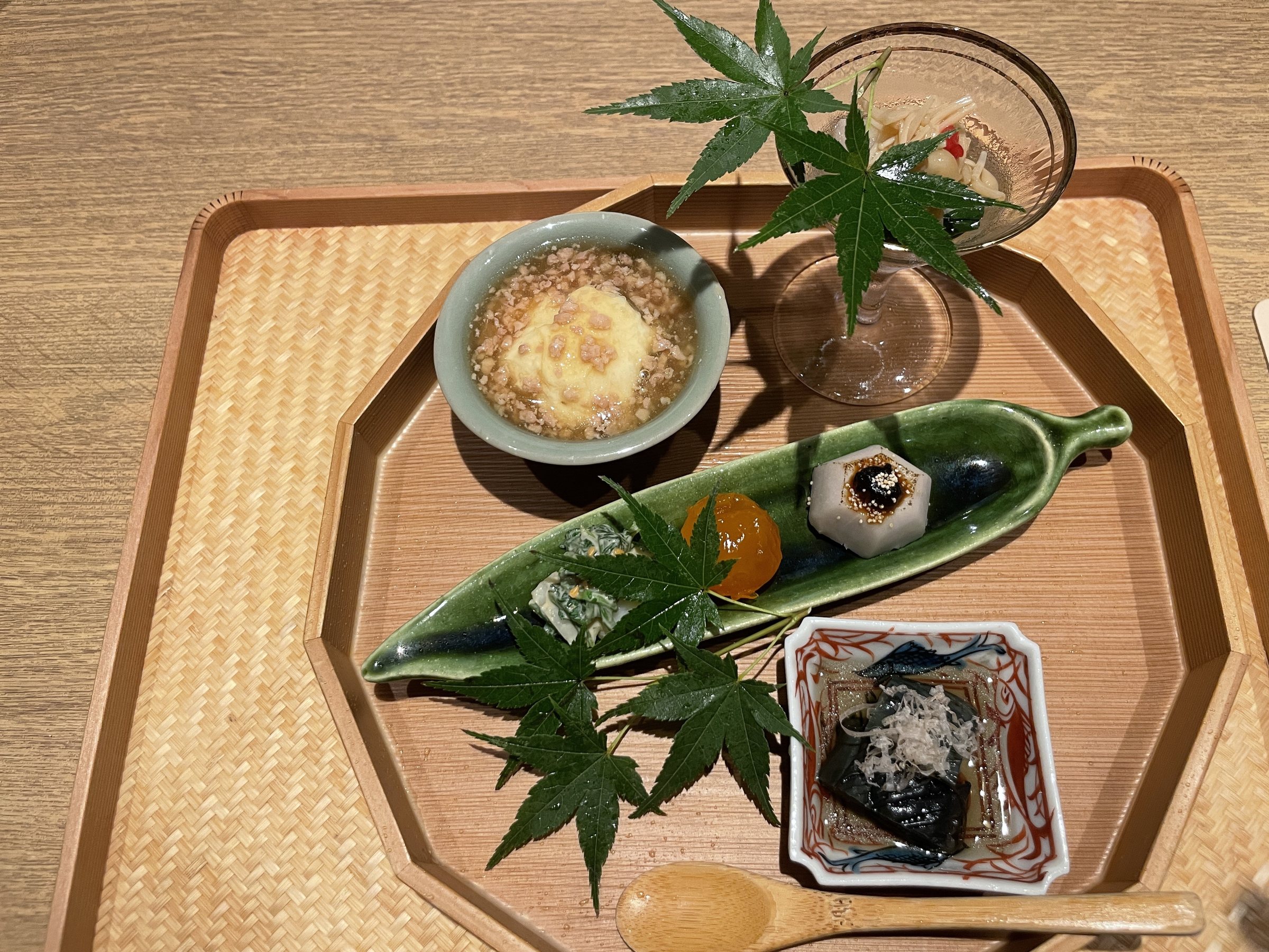 The hassun on a wooden tray dressed with maple leaves: a bamboo-leaf dish, small bowls and a glass cup.
