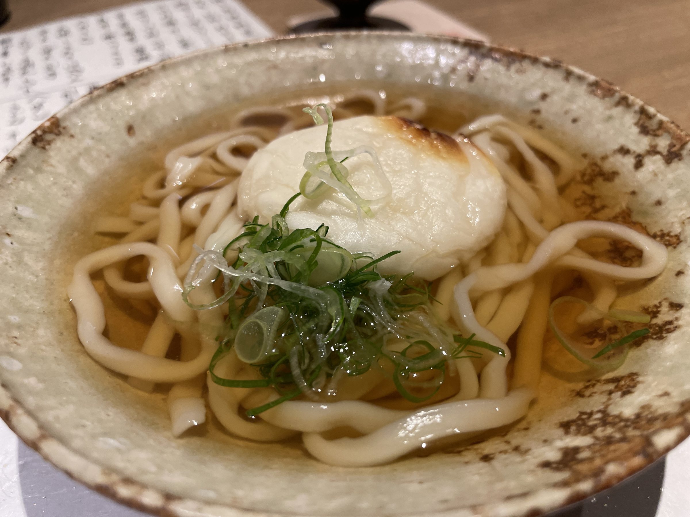 A small bowl of udon in clear broth, topped with toasted mochi and spring onion.
