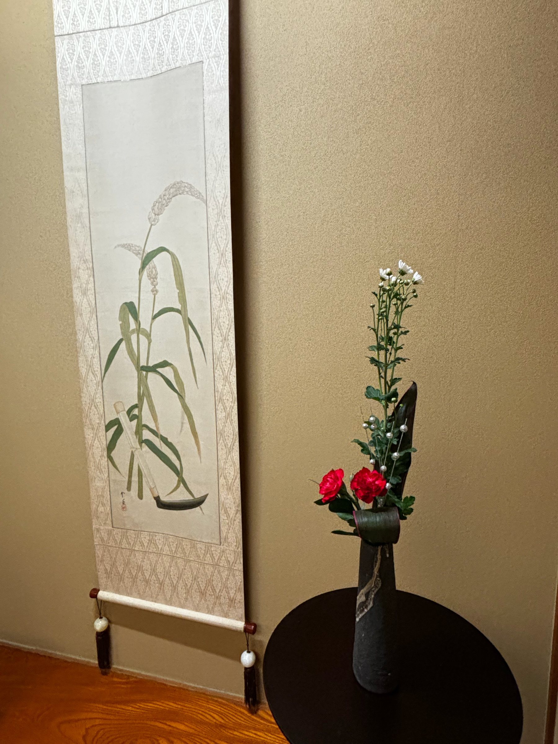 Red and white flowers and small white chrysanthemums before a scroll painted with leaves.