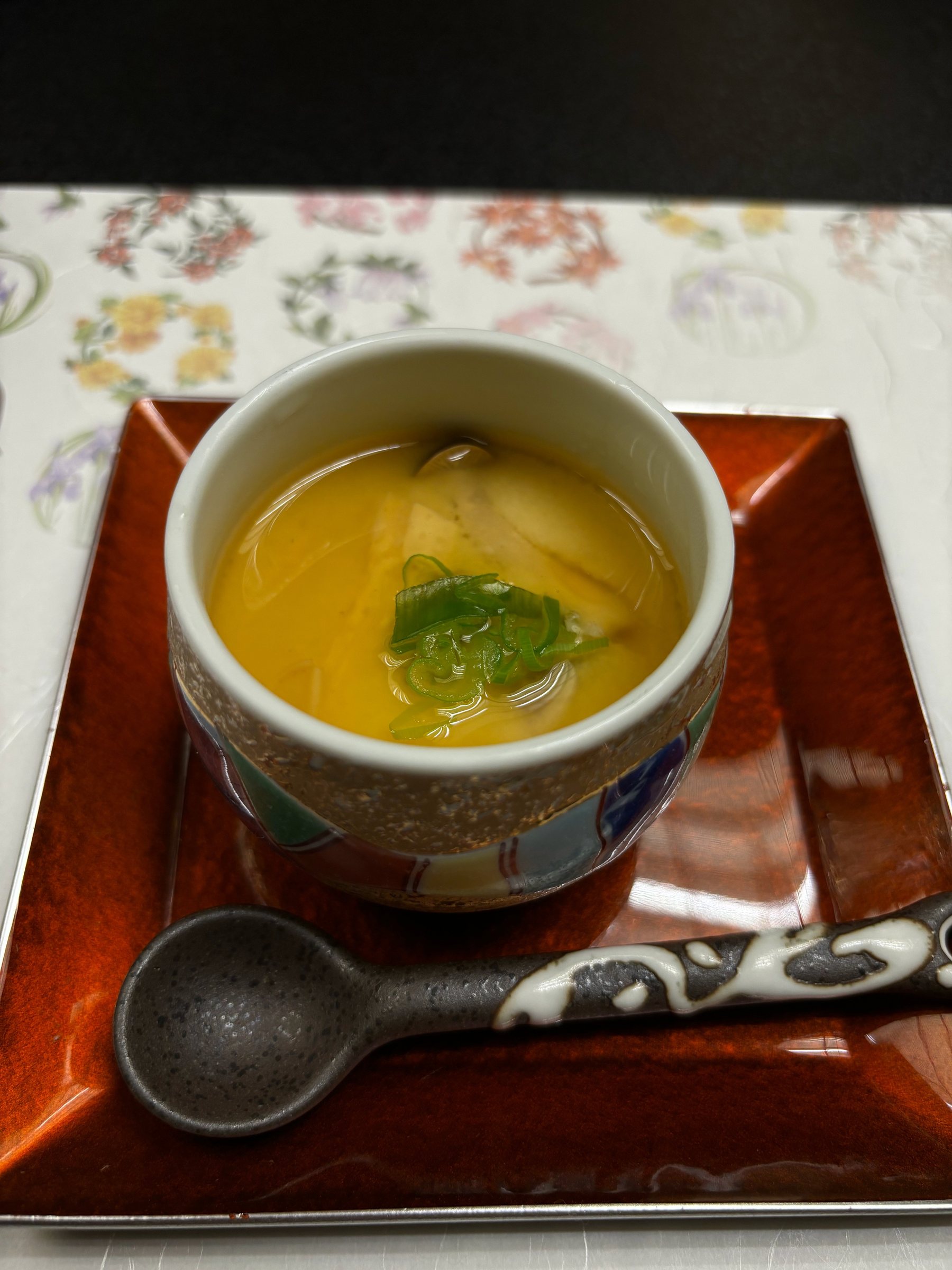 A covered cup glazed with amber sauce and topped with green onion, with a ceramic spoon.